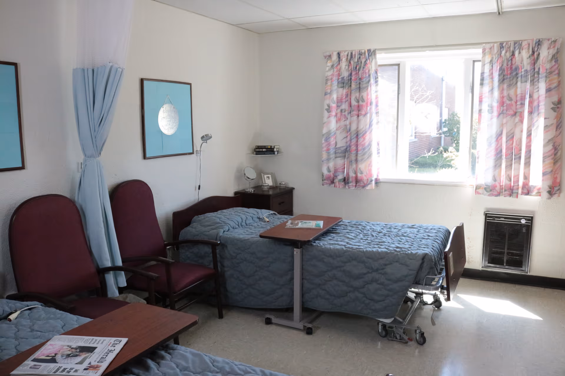 Shared patient bedroom with two hospital beds, chairs, bedside tables, and a sunlit window with floral curtains.