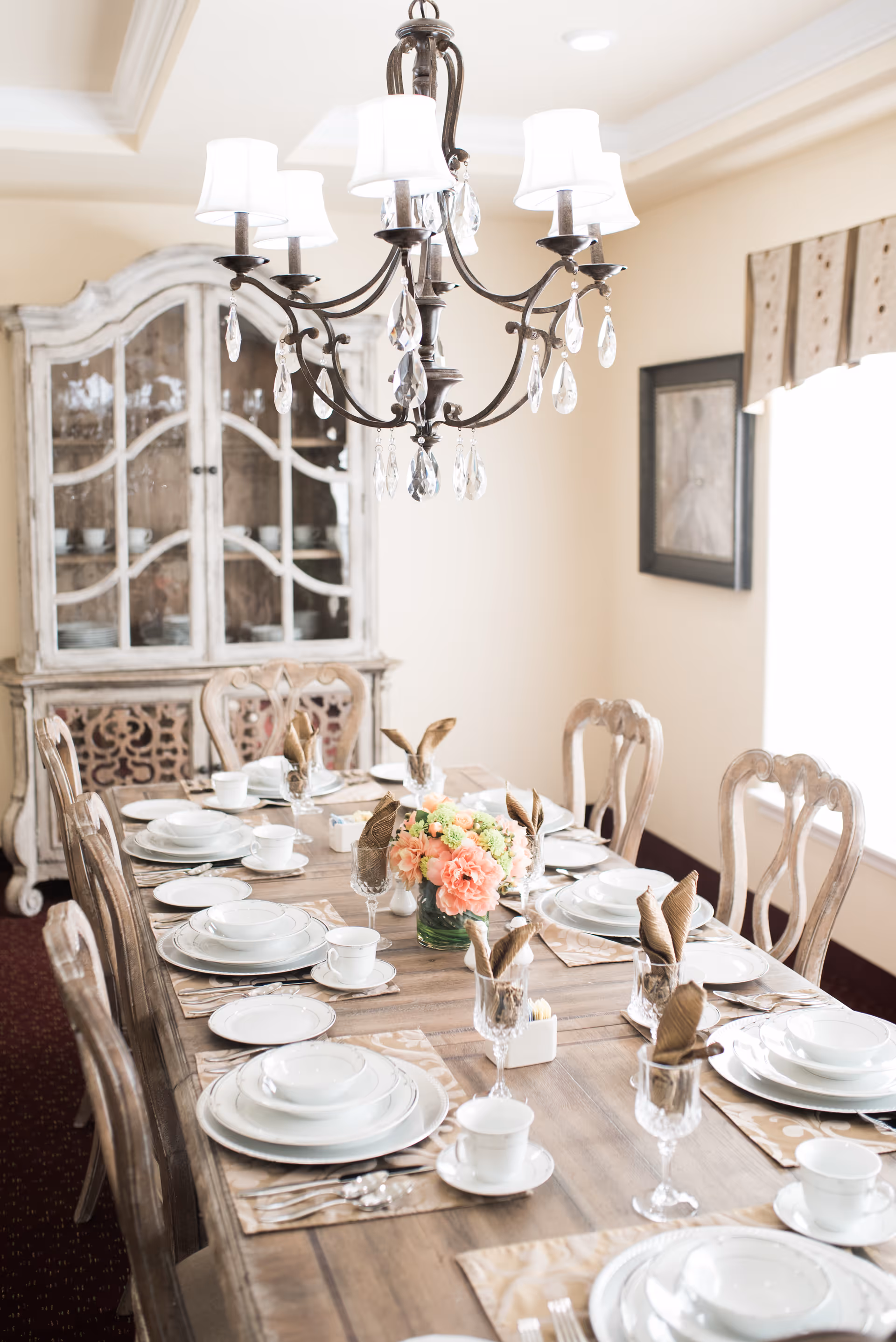 A beautifully set dining table with white plates, cups, and silverware arranged on beige placemats. Each place setting has a folded brown napkin in a glass. A floral centerpiece with pink and green flowers is in the middle of the wooden table. In the background, there is a vintage-style glass cabinet with dishes inside and a framed picture on the wall. A chandelier with white lampshades and hanging crystals is above the table.