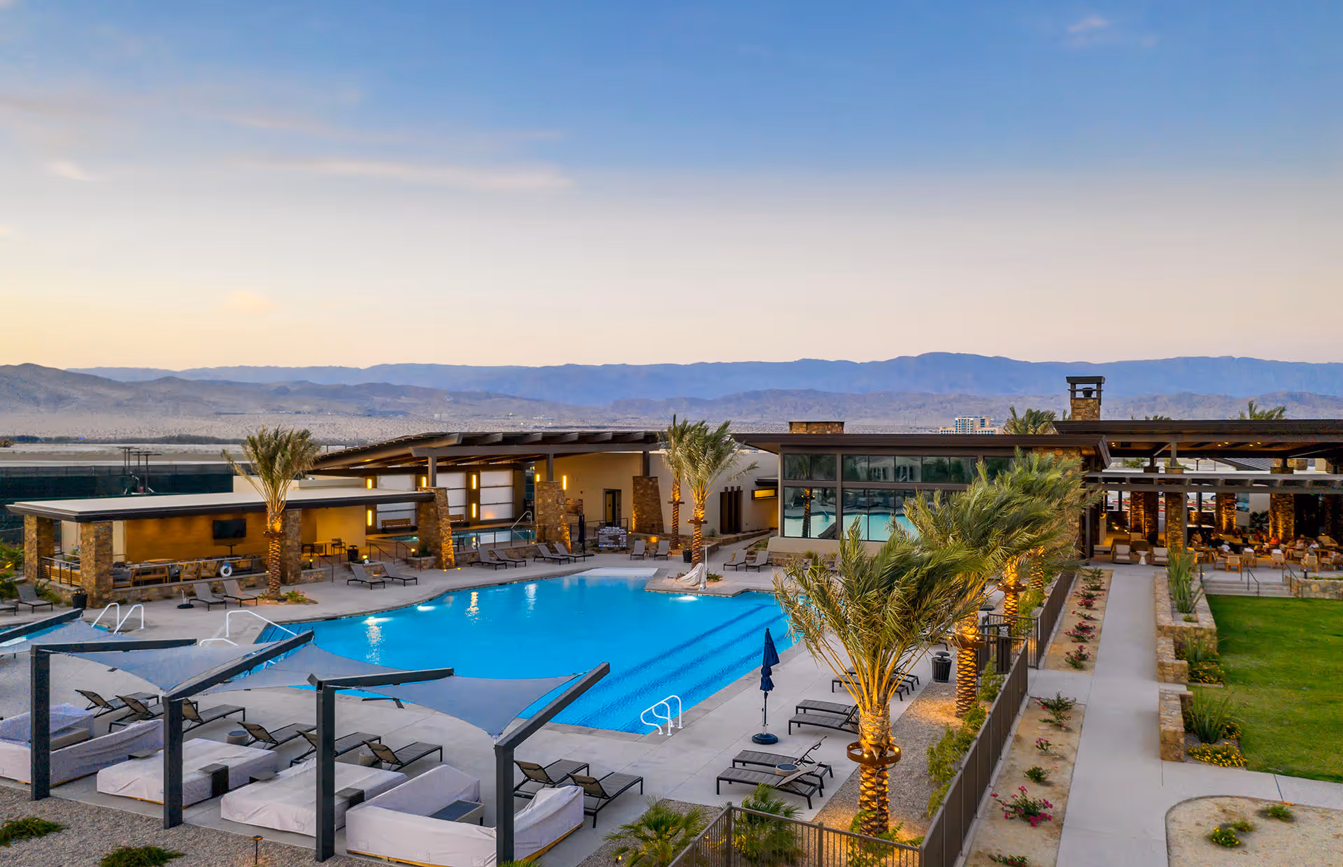 Outdoor swimming pool area at Del Webb Rancho Mirage 55+ Retirement Community with lounge chairs, shaded cabanas, palm trees, and surrounding buildings under a clear sky with mountains in the background.