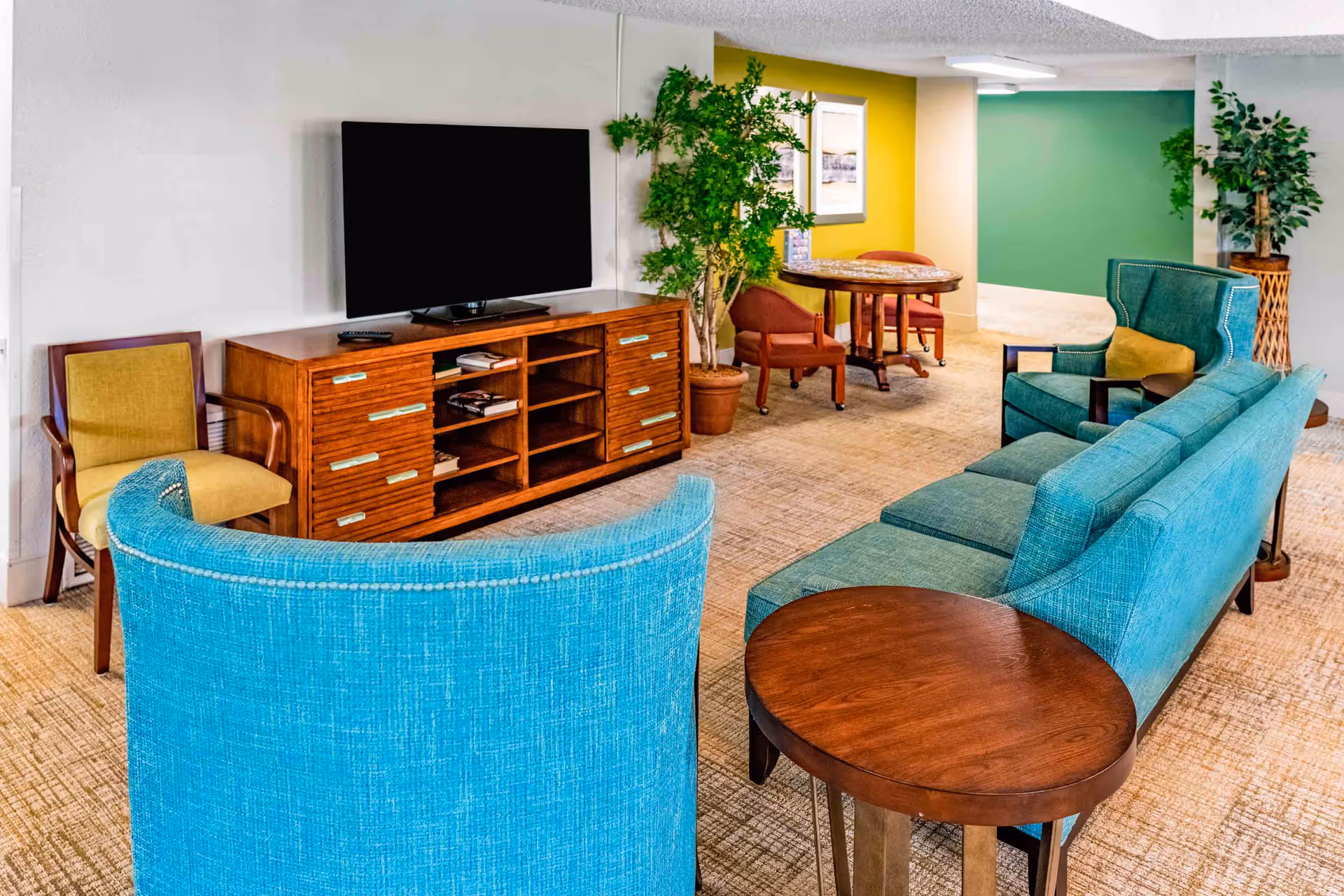 A cozy living room area with a large flat-screen TV on a wooden media console, a curved turquoise sofa, a matching turquoise armchair with a yellow cushion, a wooden side table, and additional seating including a yellow chair and two red chairs around a small round table. The walls are painted in white, yellow, and green, and there are potted plants adding greenery to the space.