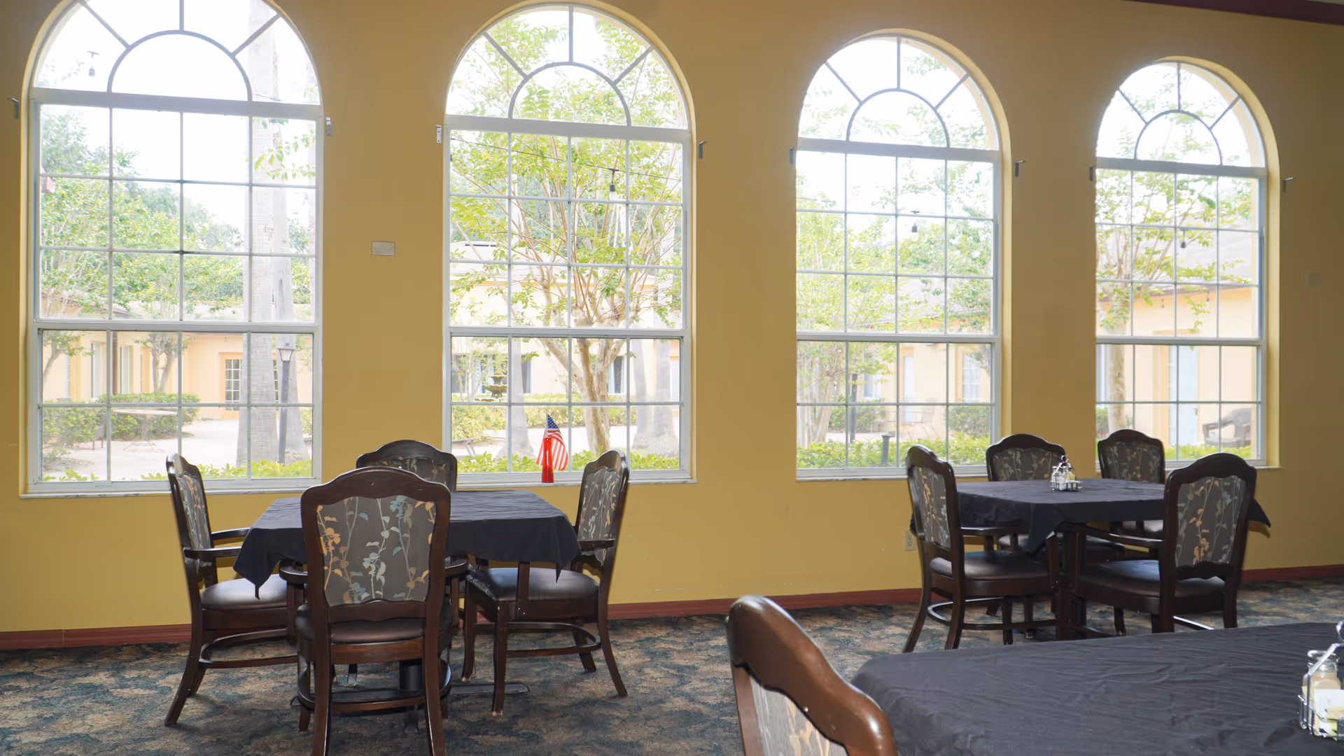 Interior dining area with multiple tables covered in black tablecloths and wooden chairs with floral upholstery. Large arched windows provide a view of trees and a courtyard outside.