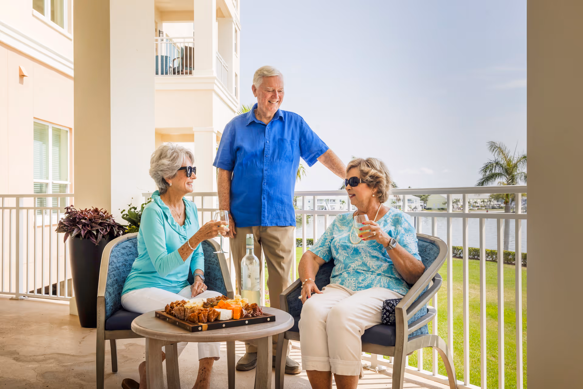 Two elderly women sitting on a balcony with a man standing between them. They are enjoying drinks and a tray of snacks on a small round table. The balcony overlooks a green lawn and a body of water with palm trees in the background.