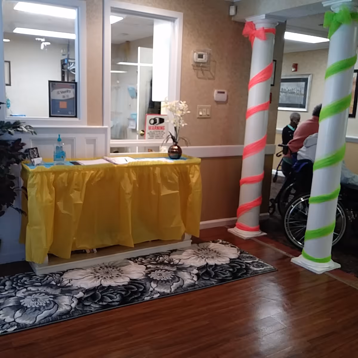 Interior view of a senior living facility reception area with a table covered in a yellow cloth, a vase with white flowers, and hand sanitizer. Two white columns wrapped with neon pink and green ribbons are visible. In the background, elderly individuals are seated, one in a wheelchair.