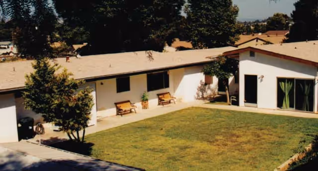 Exterior view of a single-story building with a beige roof and white walls surrounding a grassy courtyard. There are two wooden benches and some potted plants along the building walls. Trees and other houses are visible in the background under a clear sky.