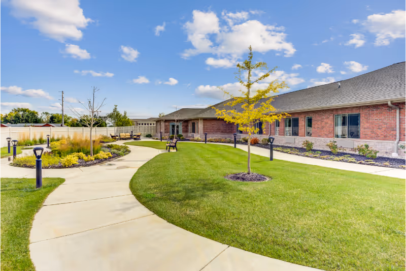 Outdoor garden area at Lakewood Senior Living with a curved concrete walkway, green grass, young trees, benches, and a brick building in the background under a partly cloudy blue sky.
