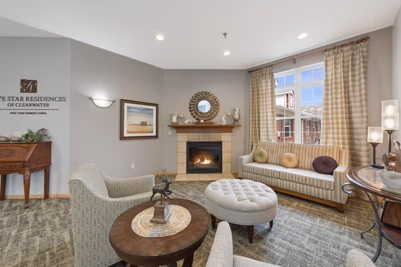 A cozy living room area in Five Star Residences of Clearwater featuring a fireplace with decorative items on the mantel, a striped sofa with round cushions, two upholstered armchairs, a round wooden coffee table with a decorative sculpture, an ottoman, a window with beige curtains, and a small wooden desk against the wall with the facility's name displayed above it.