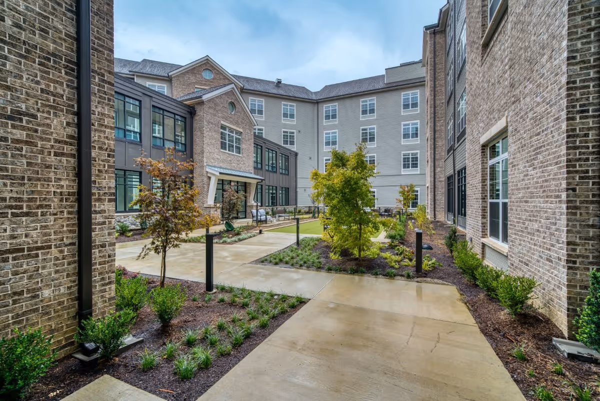 Outdoor courtyard area of a senior living facility with paved walkways, landscaped garden beds, small trees, and shrubs surrounded by multi-story brick and siding buildings under a cloudy sky.