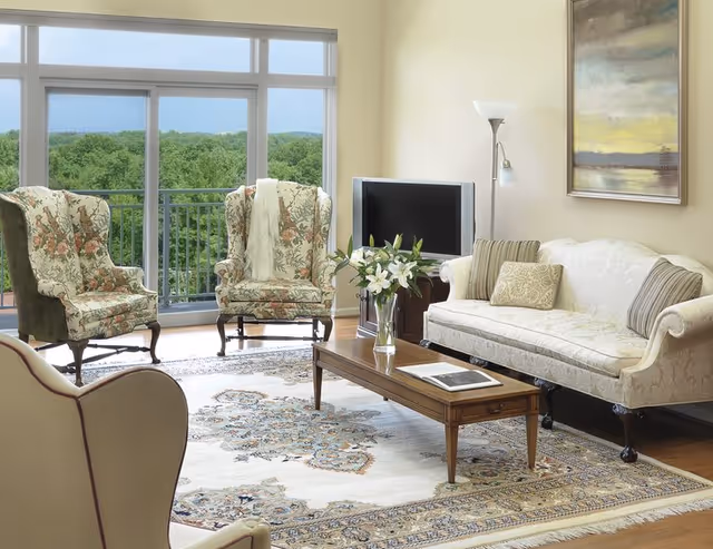 Sunlit living room with floral wingback chairs, a cream sofa, wooden coffee table with flowers, and large windows overlooking trees.