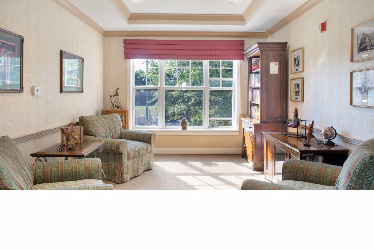 A cozy living room with three patterned armchairs arranged around a wooden side table and a wooden cabinet filled with books. The room has a large window with a red valance, allowing natural light to brighten the space. The walls are decorated with framed pictures and light-colored wallpaper.