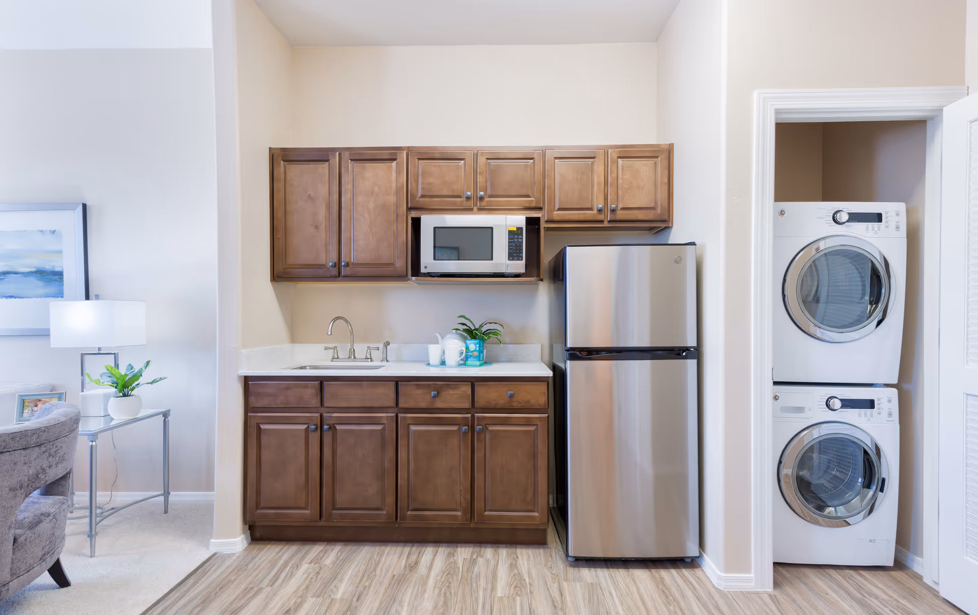 A compact kitchen area with wooden cabinets, a stainless steel refrigerator, a microwave, and a sink. To the right, there is a small laundry closet with a stacked washer and dryer. Part of a living room with a gray chair, a side table with a lamp, and a framed picture is visible on the left.