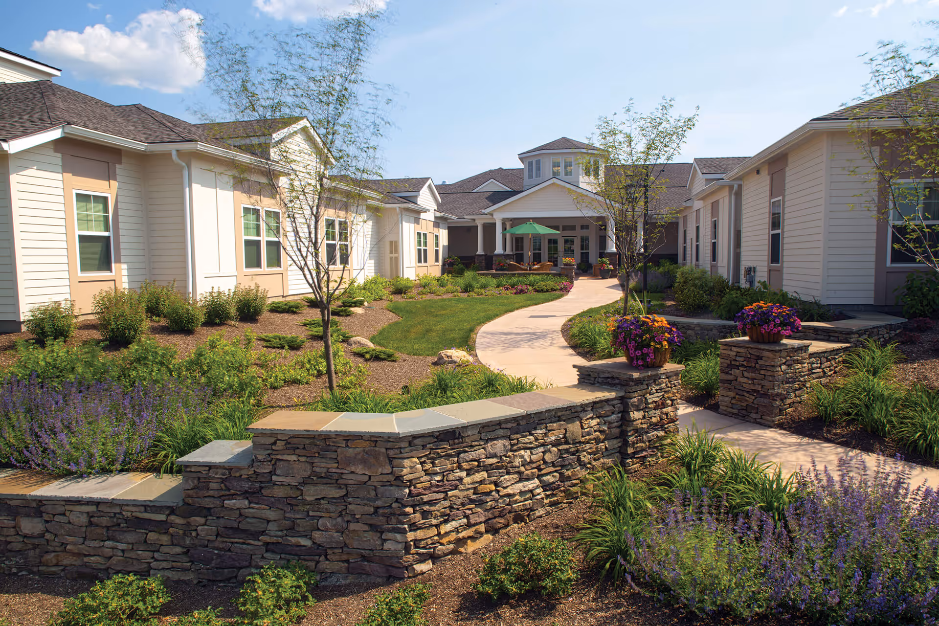 Outdoor courtyard area of Glenmere Assisted Living featuring a winding paved walkway, stone walls with flower pots, landscaped gardens with shrubs and small trees, and a building with beige siding and multiple windows under a blue sky with some clouds.
