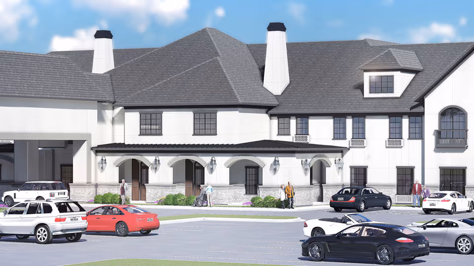 Exterior view of a large, two-story senior living facility building with white walls and gray roof. Several cars are parked in front of the building, and a few people are walking near the entrance. The sky is blue with some clouds.
