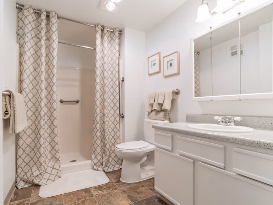 A clean bathroom with a walk-in shower enclosed by patterned curtains, a white toilet with folded towels on the tank, a countertop with a sink, and a large mirror above the sink. The floor has brown tiles and the walls are white with two framed pictures above the toilet.