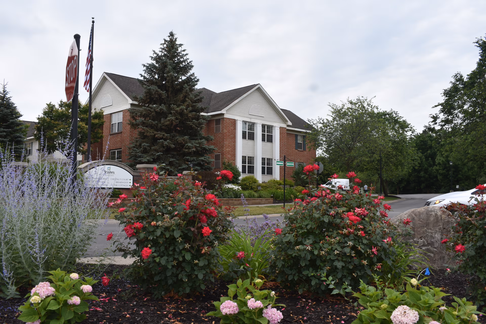 Exterior view of a two-story brick and white siding building with large windows, surrounded by trees and colorful flower bushes in the foreground. A stop sign and American flag are visible near the building entrance.
