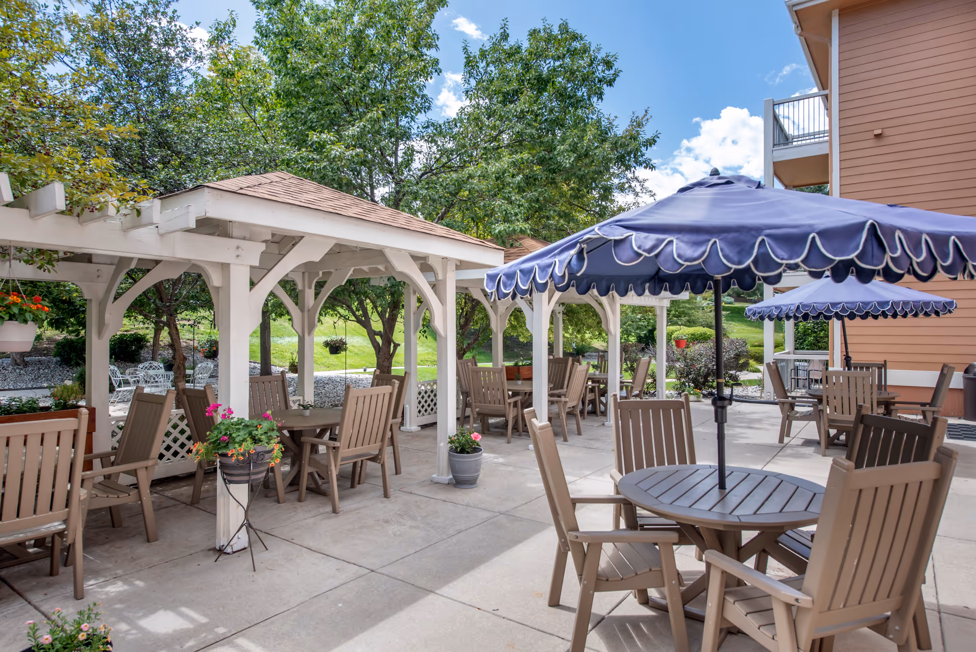Outdoor patio area at Brookdale Vista Grande featuring multiple tables with brown chairs and blue umbrellas. There are white wooden pergolas providing shade, potted plants, and green trees in the background under a partly cloudy sky.