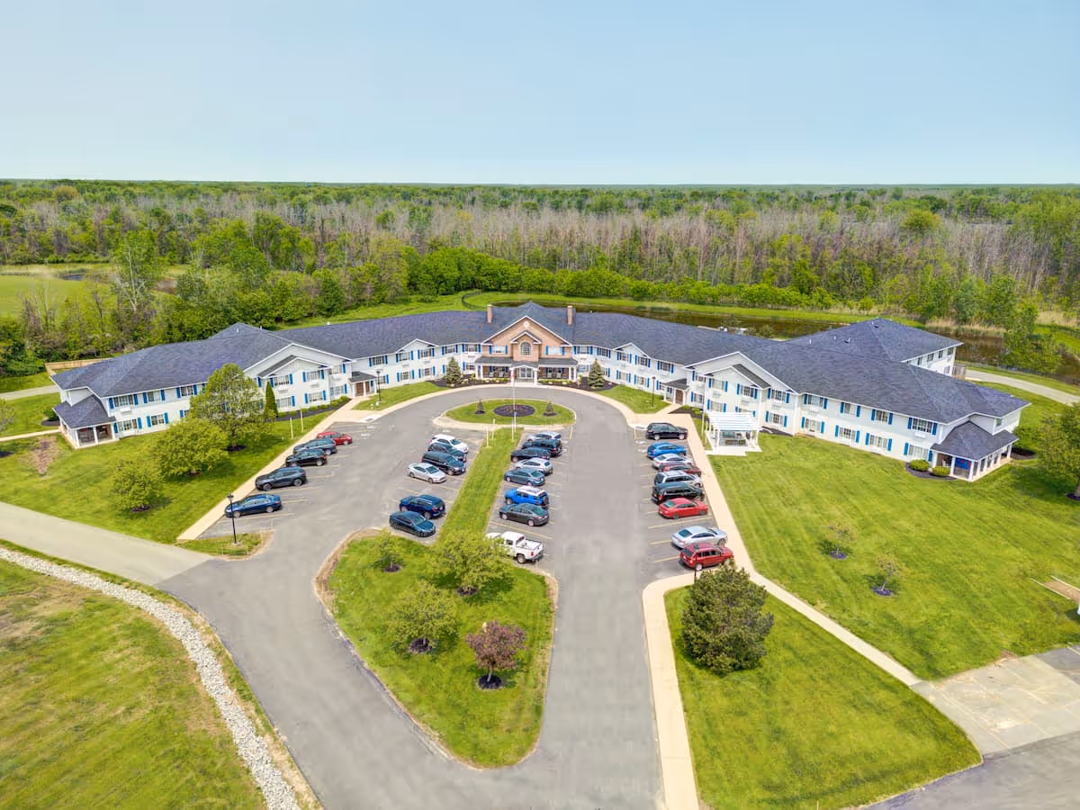 Aerial view of Evergreen Place, a large senior living facility with a U-shaped building surrounded by green lawns and trees. The building has a dark roof and white exterior with blue window shutters. There is a parking lot with several cars in front of the main entrance, and a circular driveway with landscaped islands.