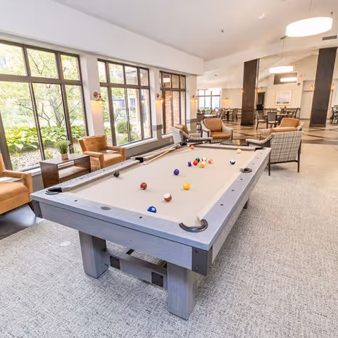 A bright and spacious common area featuring a pool table with billiard balls arranged on it, surrounded by comfortable armchairs and large windows letting in natural light. The room has a modern and inviting atmosphere with additional seating and tables in the background.