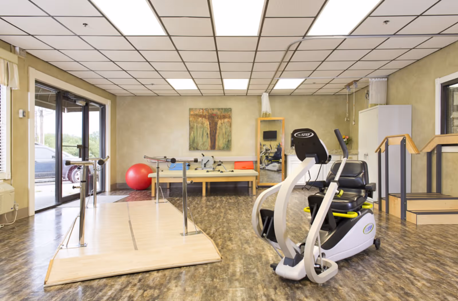 Bright interior therapy/exercise room with parallel bars, a recumbent exercise bike, a red exercise ball and benches near sliding glass doors.