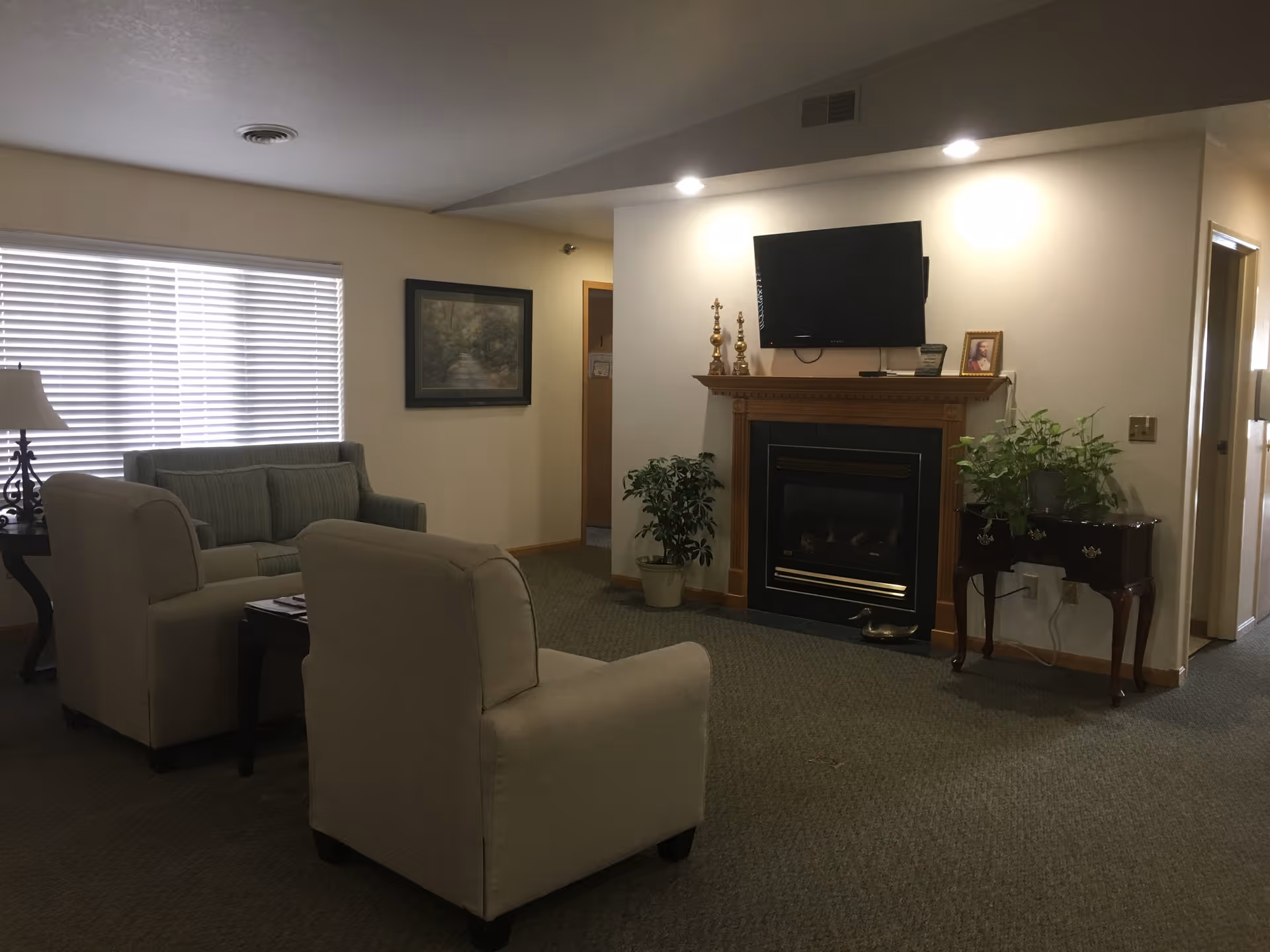 Cozy common room with upholstered chairs and a sofa by a window, a fireplace with a TV mounted above, and potted plants.