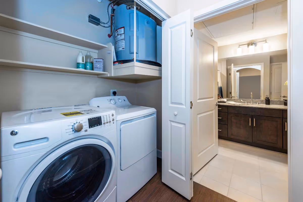 Laundry area with a front-loading washer and dryer and water heater, with a door opening into a bathroom with a double-sink vanity.