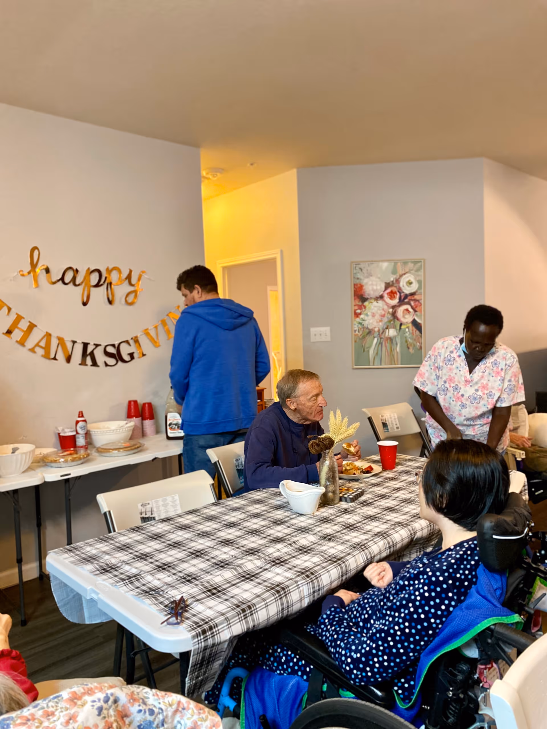 A group of people gathered in a room decorated with a 'happy THANKSGIVING' banner on the wall. There is a table covered with a plaid tablecloth, with some food and drinks on it. One person in a wheelchair is seated at the table, while another person is sitting and eating. A person in a blue hoodie is standing near a buffet table with food and condiments, and another person in a floral shirt is standing nearby. The room has light-colored walls and a floral painting hanging on one wall.