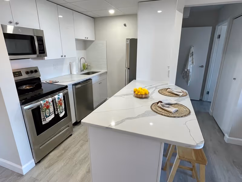 Modern kitchen with white cabinets, stainless steel appliances including a microwave, stove, dishwasher, and refrigerator. A white marble countertop island with two place settings and a bowl of lemons is in the foreground. Light wood flooring and neutral walls complete the space.