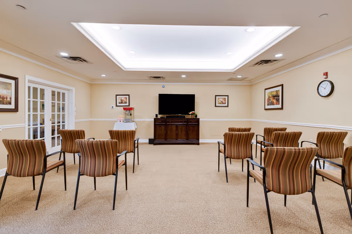 A meeting or activity room with several striped upholstered chairs arranged in rows facing a flat-screen TV mounted on a wooden cabinet. The room has beige walls with framed artwork, a clock on the wall, and a popcorn machine on a small table covered with a white cloth. The ceiling features recessed lighting and a large central light panel.