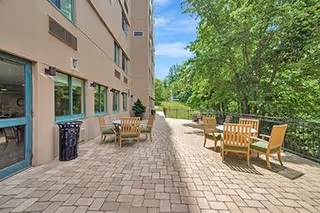 Outdoor patio area at Chestnut Ridge Wallingford with several wooden tables and chairs arranged on a paved surface next to a building. The area is bordered by a black metal railing and surrounded by green trees and grass under a clear blue sky.