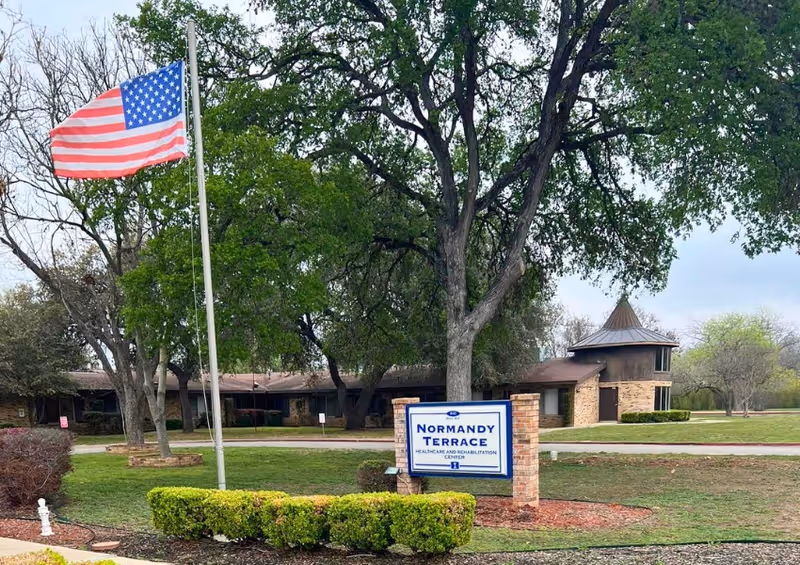 Outdoor view of Normandy Terrace Healthcare and Rehabilitation Center with a large American flag on a flagpole, a sign displaying the facility's name, and surrounding trees and greenery.