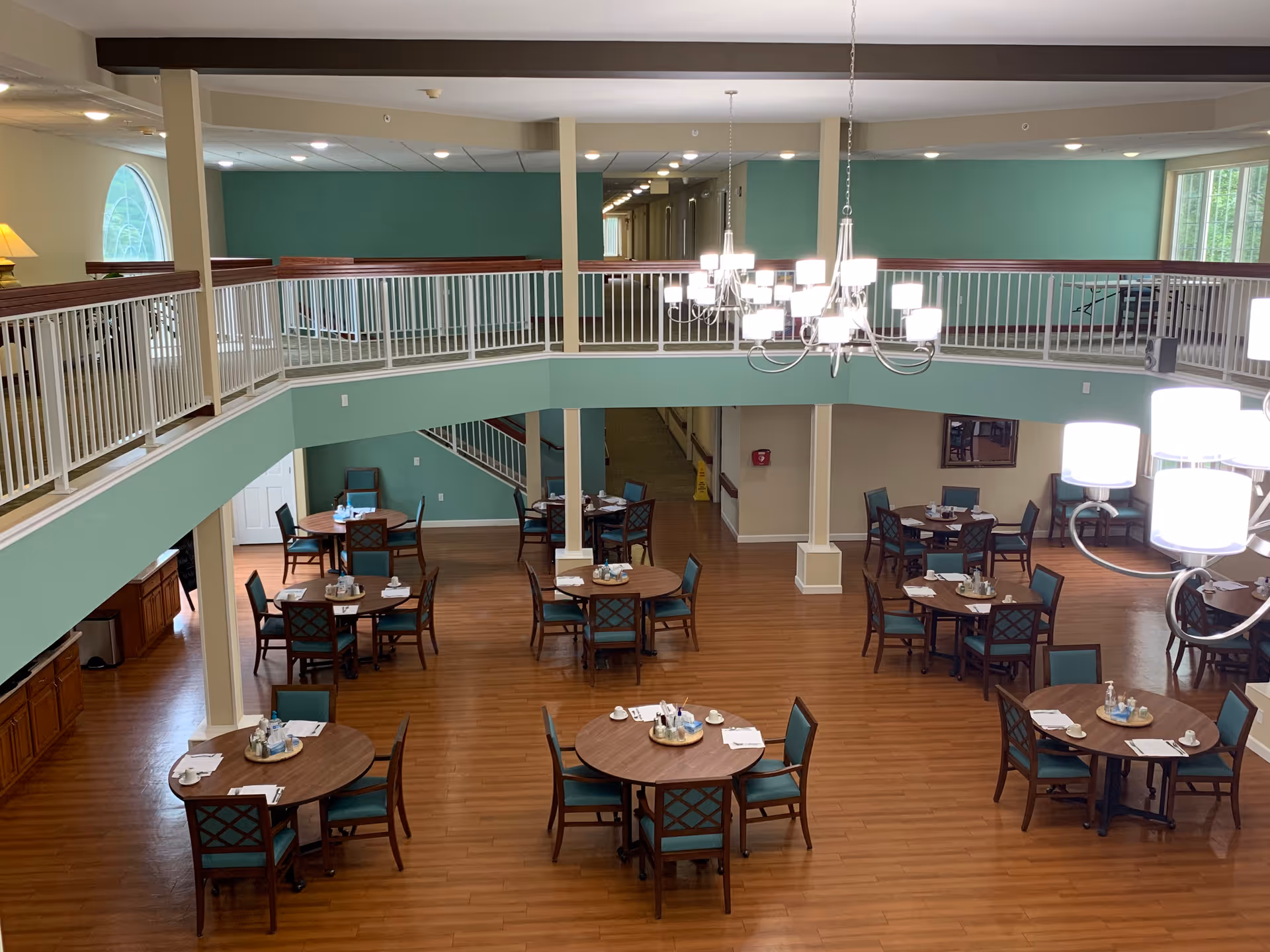 Interior view of a senior living facility dining room with multiple round wooden tables and teal cushioned chairs arranged neatly on a wooden floor. The room features a two-story design with a white railing balcony overlooking the dining area. Modern chandeliers hang from the ceiling, providing bright lighting. The walls are painted in a soft green and beige color scheme.
