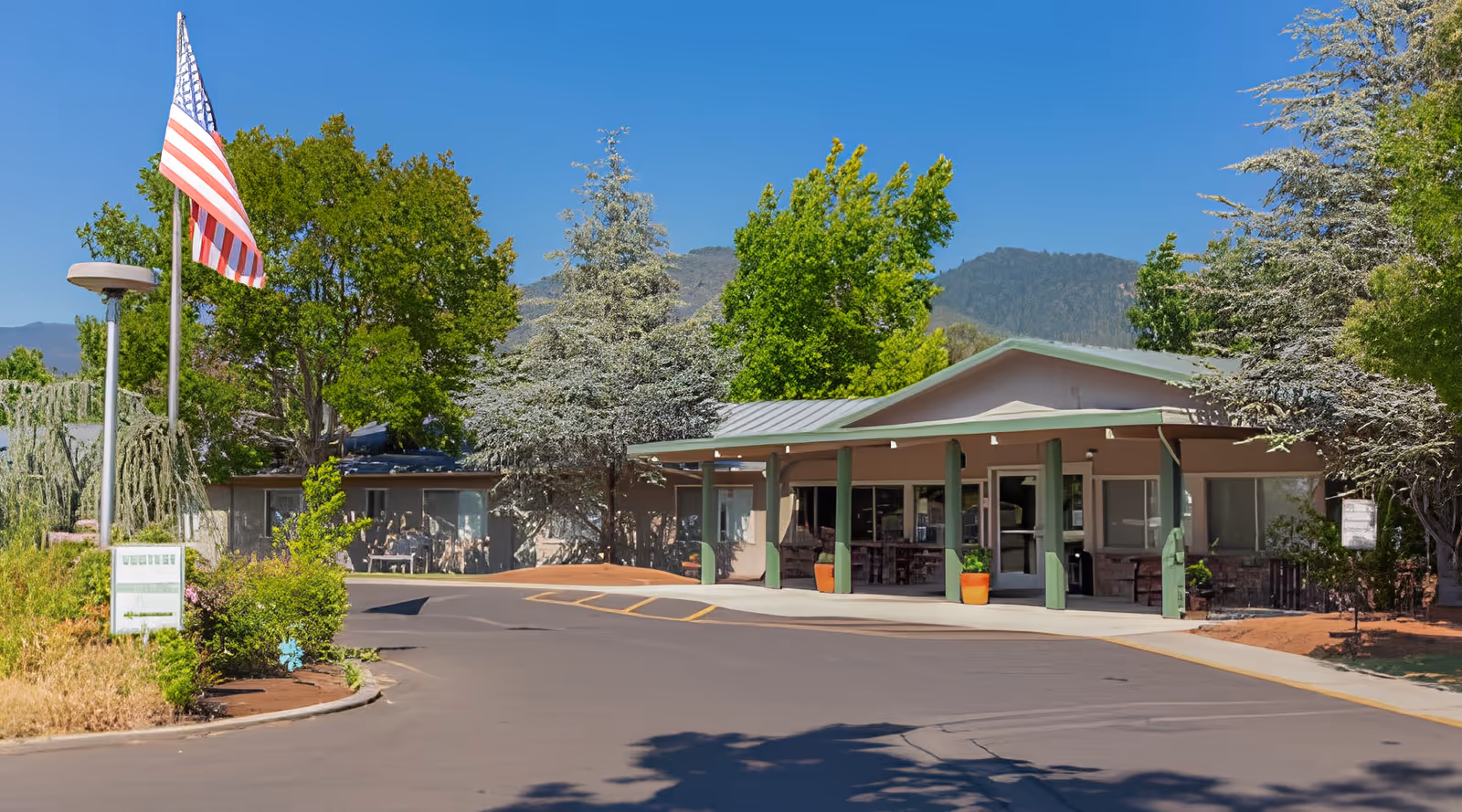 Exterior view of Royale Gardens Health & Rehabilitation Center showing the entrance with a covered porch supported by green pillars, surrounded by trees and landscaping under a clear blue sky. An American flag is flying on a flagpole near the driveway.