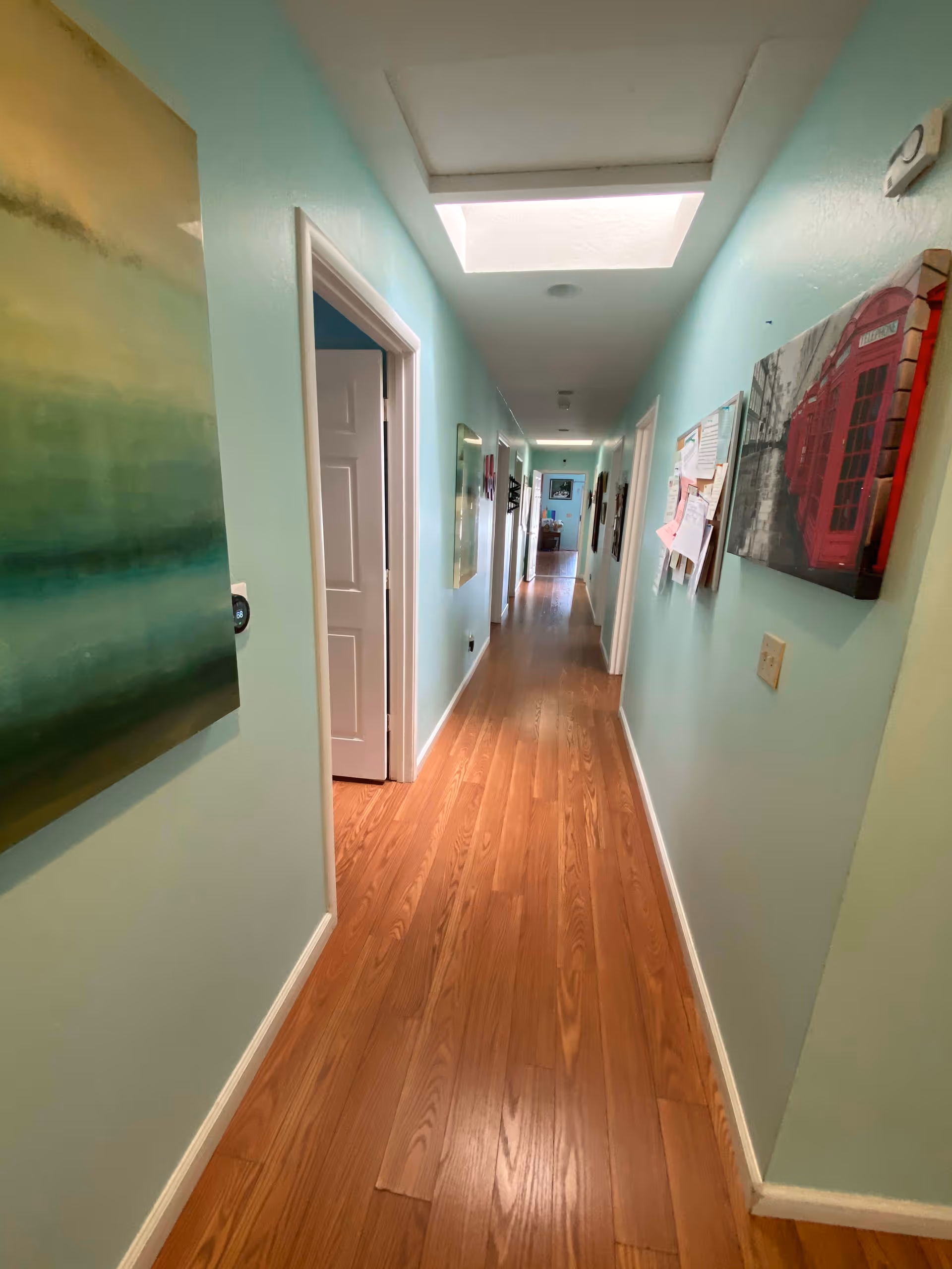 A long, narrow hallway with light blue walls and wooden flooring. The hallway is decorated with various paintings and a bulletin board on the right wall. There is a skylight on the ceiling providing natural light. Several doorways lead off the hallway, and the end of the hallway opens into another room.