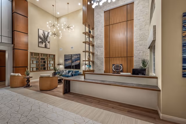 Spacious and modern senior living facility lobby with high ceilings, contemporary pendant lights, a wooden reception desk, comfortable seating area with chairs and a sofa, decorative wall art, and a mix of light stone and wood paneling on the walls.