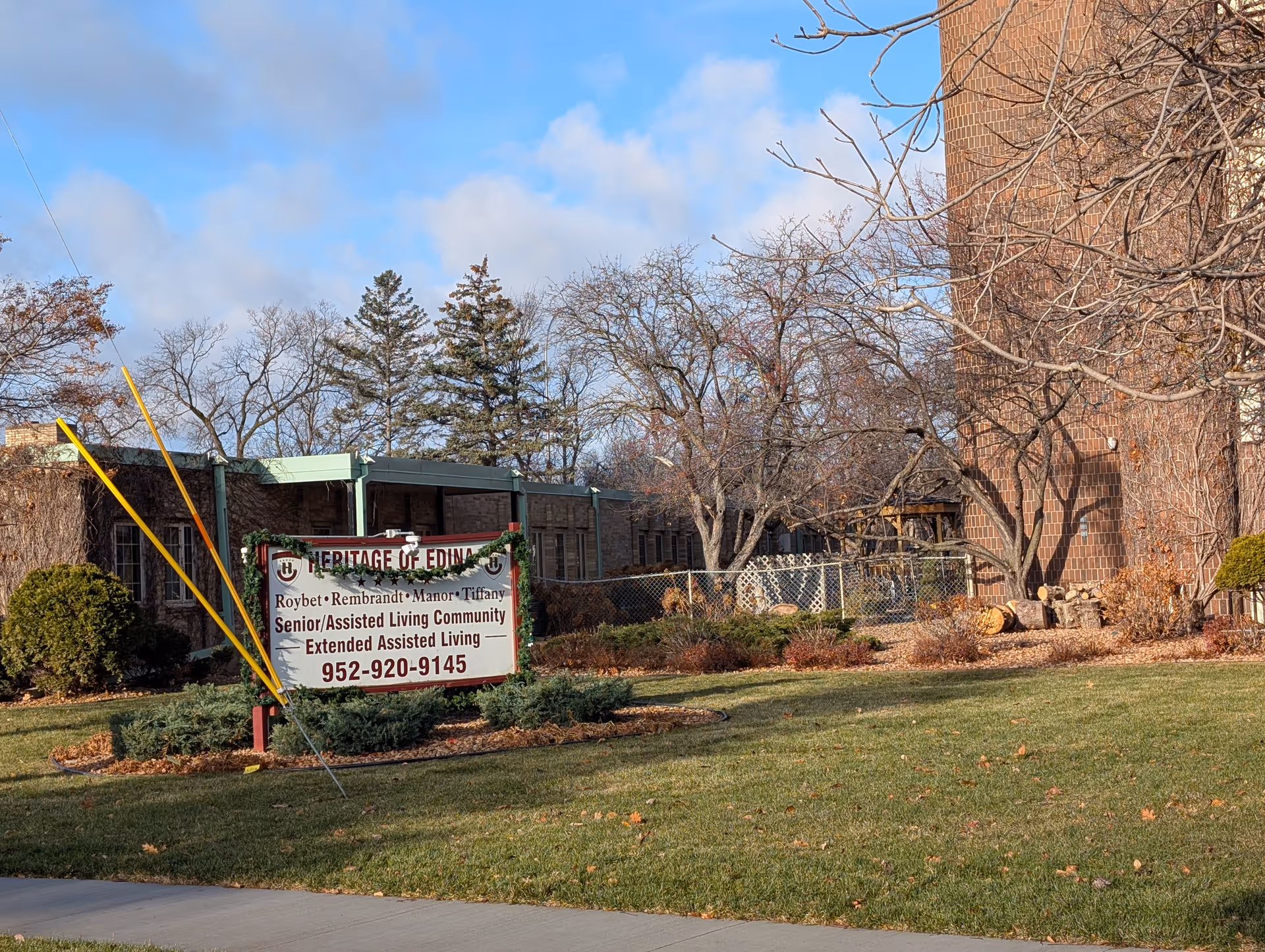Front lawn and sign for Heritage Of Edina senior living community in front of a brick building and leafless trees.