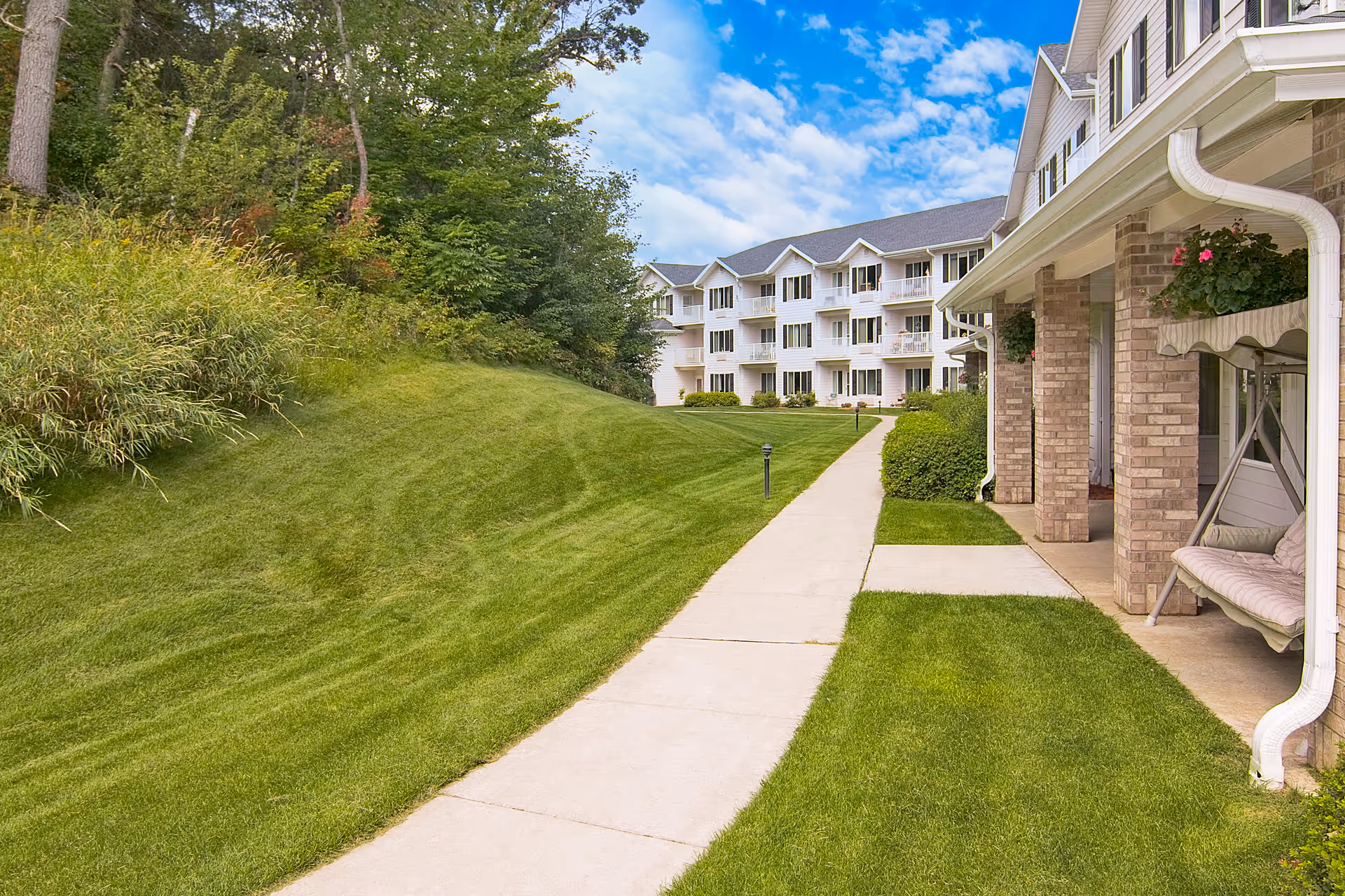 A paved walkway runs beside a manicured lawn toward a multi-story apartment building with balconies and a covered porch swing.