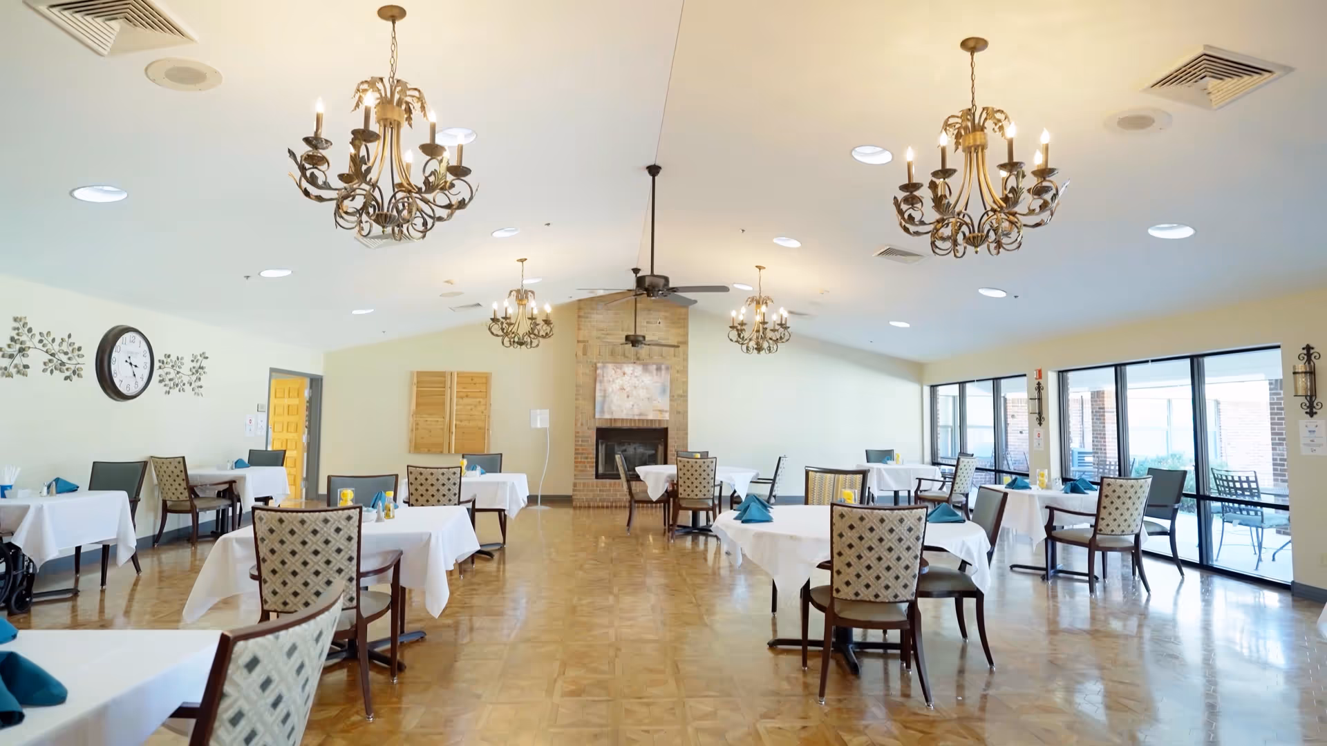 A spacious dining room with multiple tables covered in white tablecloths and set with blue napkins. The room features patterned chairs, a wooden parquet floor, decorative chandeliers hanging from the ceiling, a brick fireplace at the far end, and large windows along one wall letting in natural light.