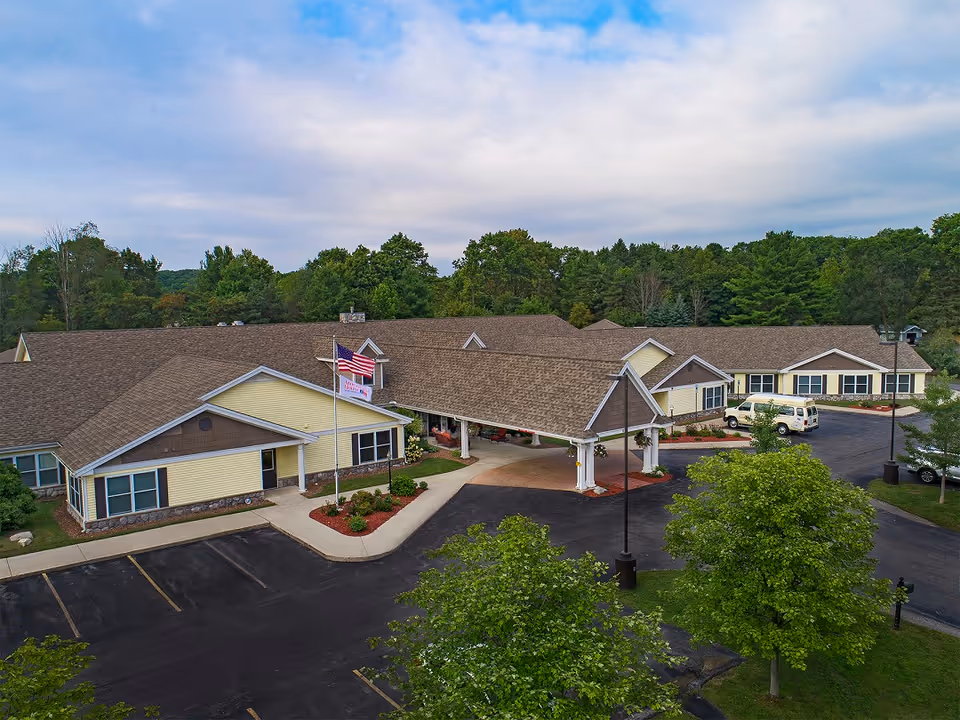 Aerial view of the front entrance of a single-story senior living building with a covered porte-cochère, American flag, parking lot and surrounding trees.