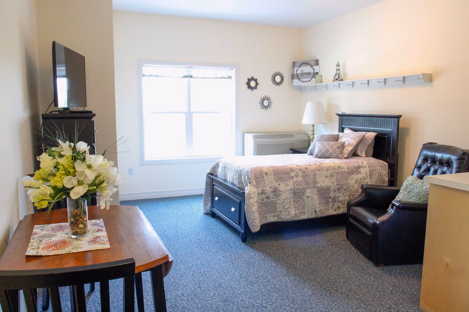A bright and cozy bedroom with a large window letting in natural light. The room features a bed with a patterned quilt, several pillows, a black leather armchair with a cushion, a wooden table with a floral centerpiece, a TV on a stand, and wall decorations including small mirrors and a shelf with decorative items.