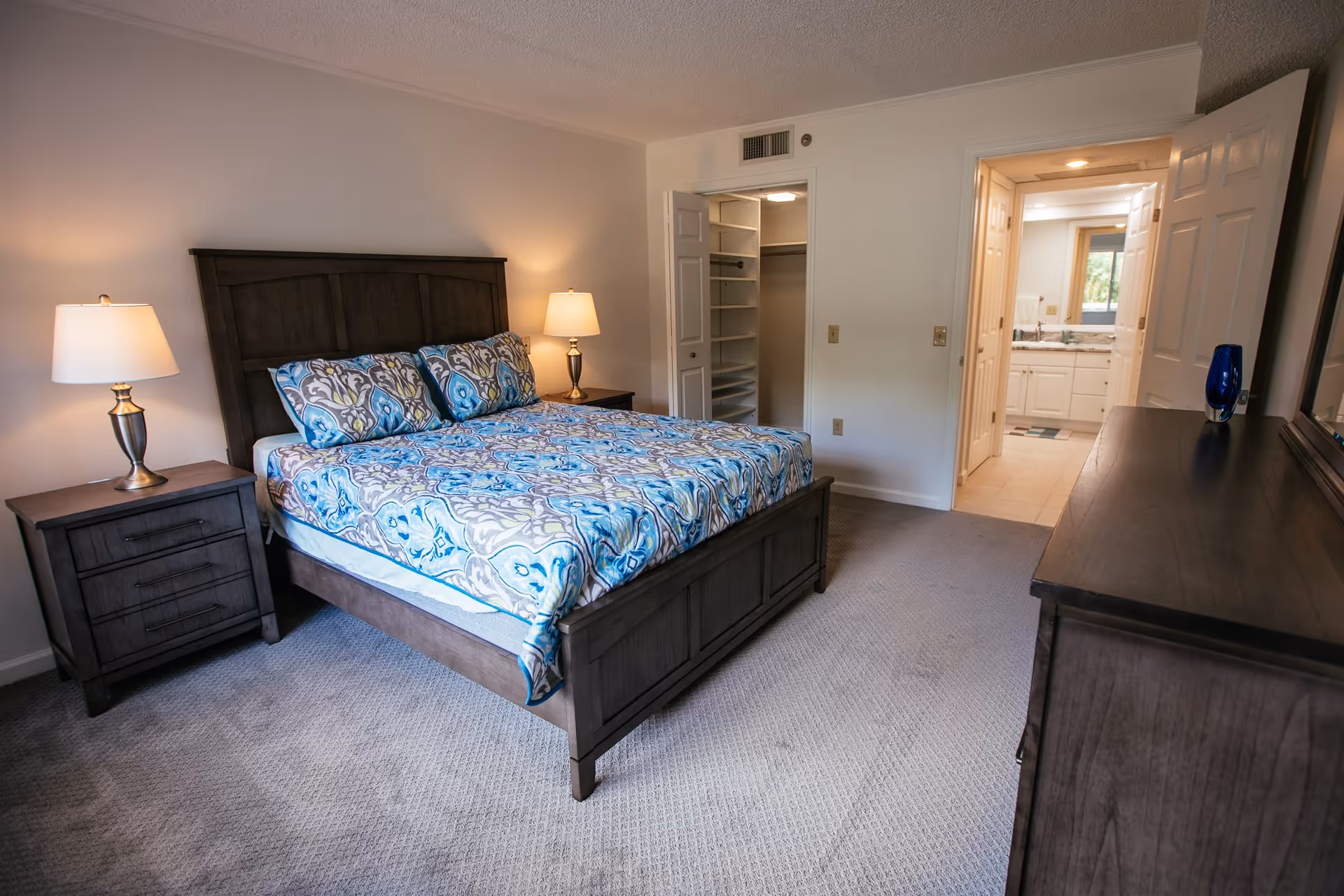 Bright furnished bedroom featuring a wooden bed with patterned bedding, two nightstands with lamps, a dresser, open closet and a view into the adjoining bathroom.