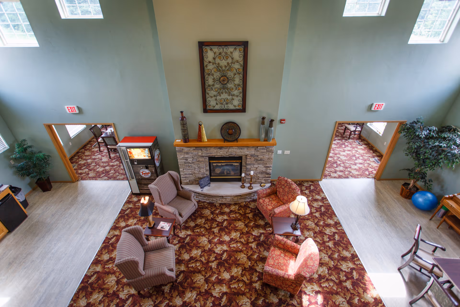 A cozy senior living facility common area with a patterned carpet, four upholstered armchairs arranged around a small table with a lamp, and a stone fireplace with decorative items on the mantel. There are two doorways leading to adjacent rooms with similar carpeting and chairs. Large windows let in natural light, and there are potted plants and a blue exercise ball in the corners.