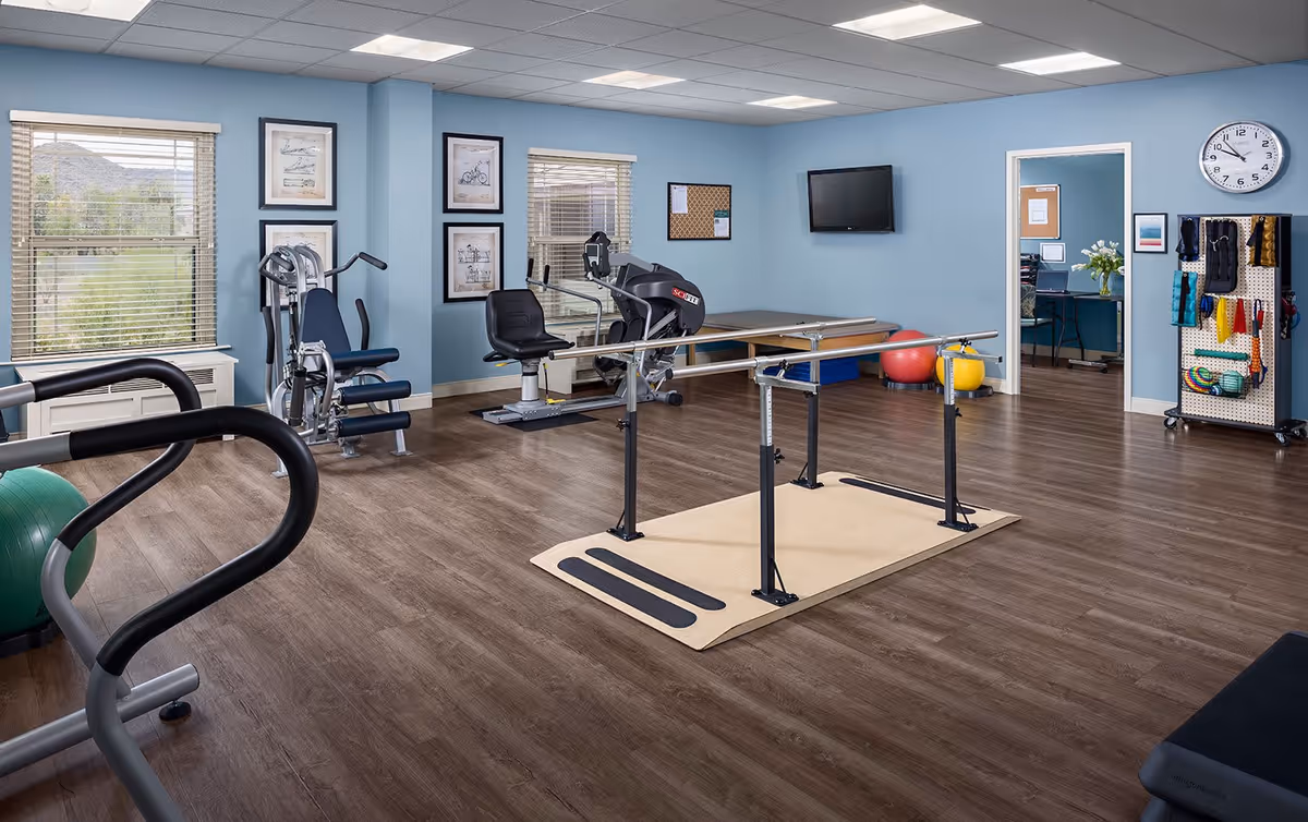 A senior living facility exercise room with light blue walls, wood flooring, and various exercise equipment including a stationary bike, weight machine, parallel bars for walking support, and exercise balls. There are two windows with blinds, framed pictures on the wall, a wall-mounted TV, a clock, and a pegboard with exercise accessories. An open doorway leads to an office area with a desk and computer.