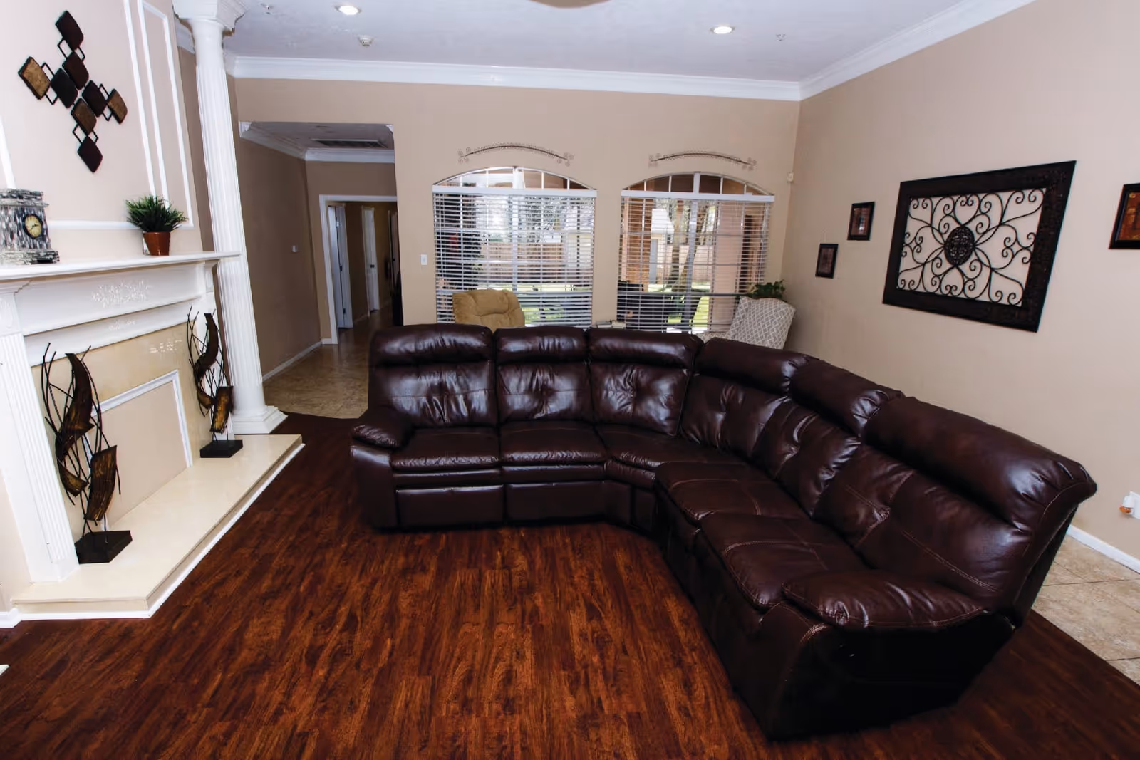 A living room with a large dark brown leather sectional sofa on wooden flooring. The room features a white fireplace with decorative items on the mantel, beige walls with framed artwork, two windows with white blinds, and a beige armchair in the corner.