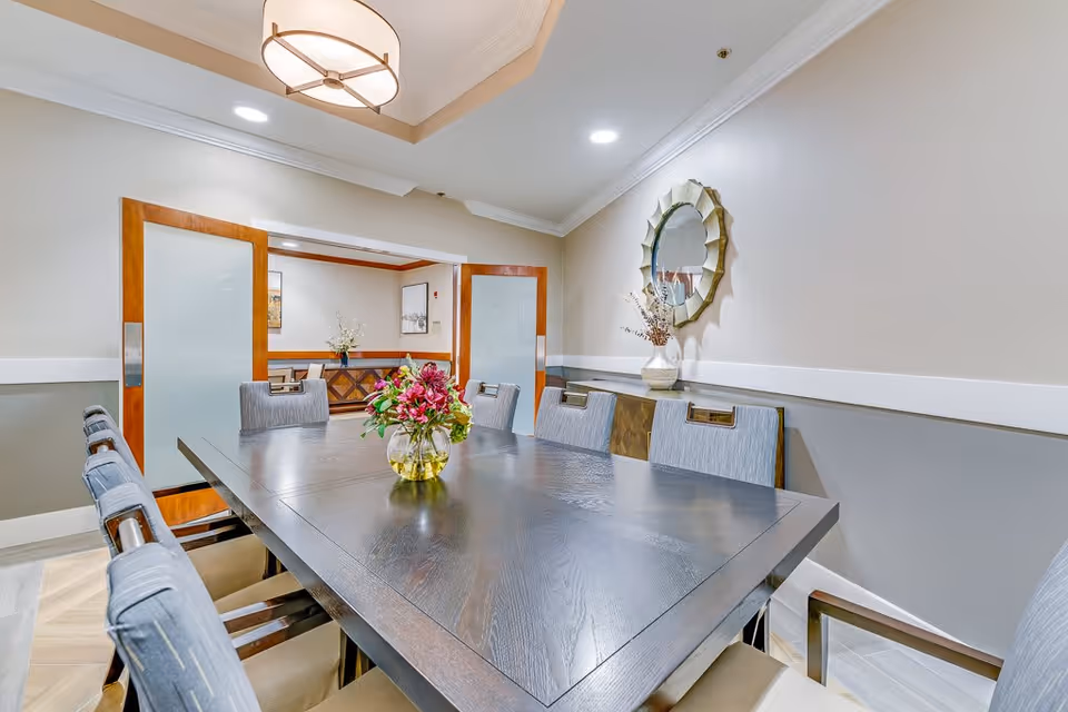 A modern dining room with a large dark wooden table surrounded by eight cushioned chairs. A vase with colorful flowers is placed in the center of the table. The room features beige walls with white trim, a decorative round mirror on one wall, and a sideboard beneath it. Two wooden sliding doors with frosted glass panels lead to an adjacent room with additional seating and artwork on the walls. The ceiling has recessed lighting and a large circular light fixture.