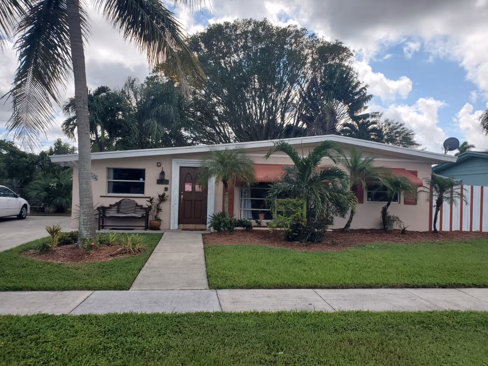 Single-story residential building with a brown front door, a bench to the left of the door, and several palm trees and shrubs in the front yard. The building has a light pink exterior with red awnings over the windows. A concrete walkway leads from the sidewalk to the front door. A white car is parked on the driveway to the left of the house, and there is a large tree behind the building under a partly cloudy sky.