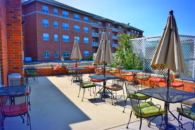 Outdoor patio area with multiple metal tables and chairs, each table shaded by a closed umbrella. Cushions in various colors are on the chairs. A brick building is visible in the background under a clear blue sky.