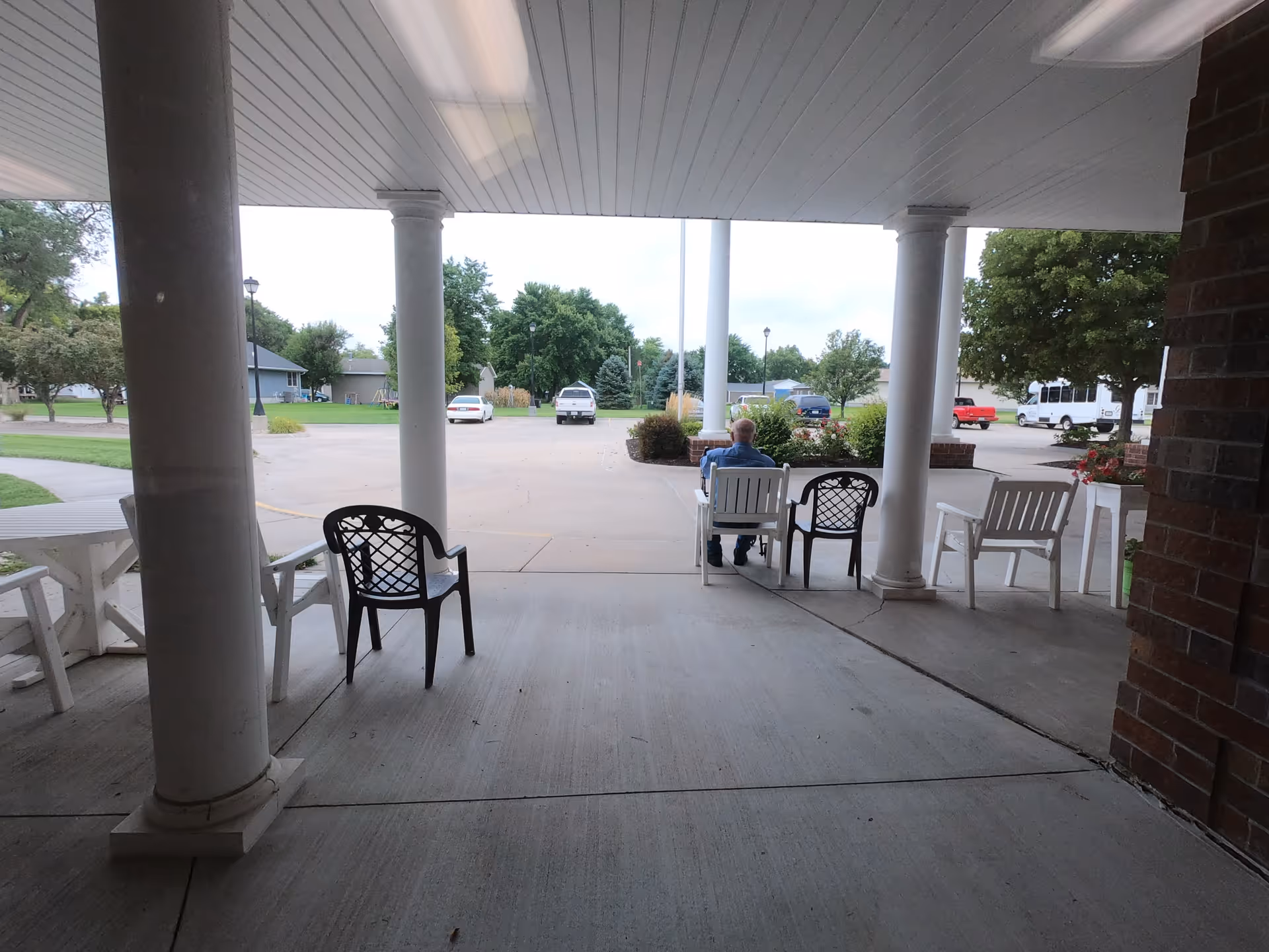 View from a covered porch area with white columns, several chairs, and a person sitting facing away looking out towards a parking lot with cars and trees in the background.