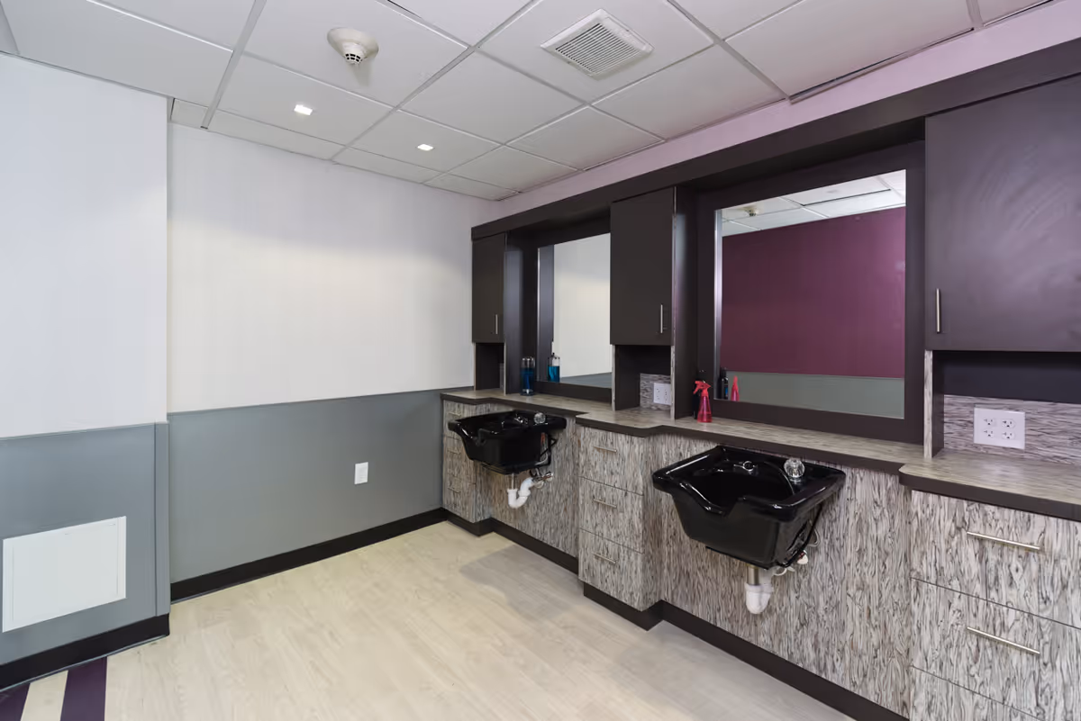 Interior view of a salon area with two black hair washing sinks mounted on a countertop with cabinets below and mirrors above. The walls are painted in light gray and purple tones, and the ceiling has recessed lighting.