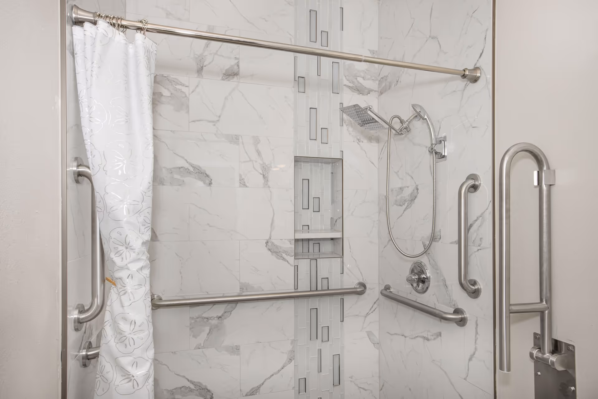 Accessible tiled shower with multiple stainless grab bars, a rainfall and handheld showerhead, built-in niche, and a patterned shower curtain.