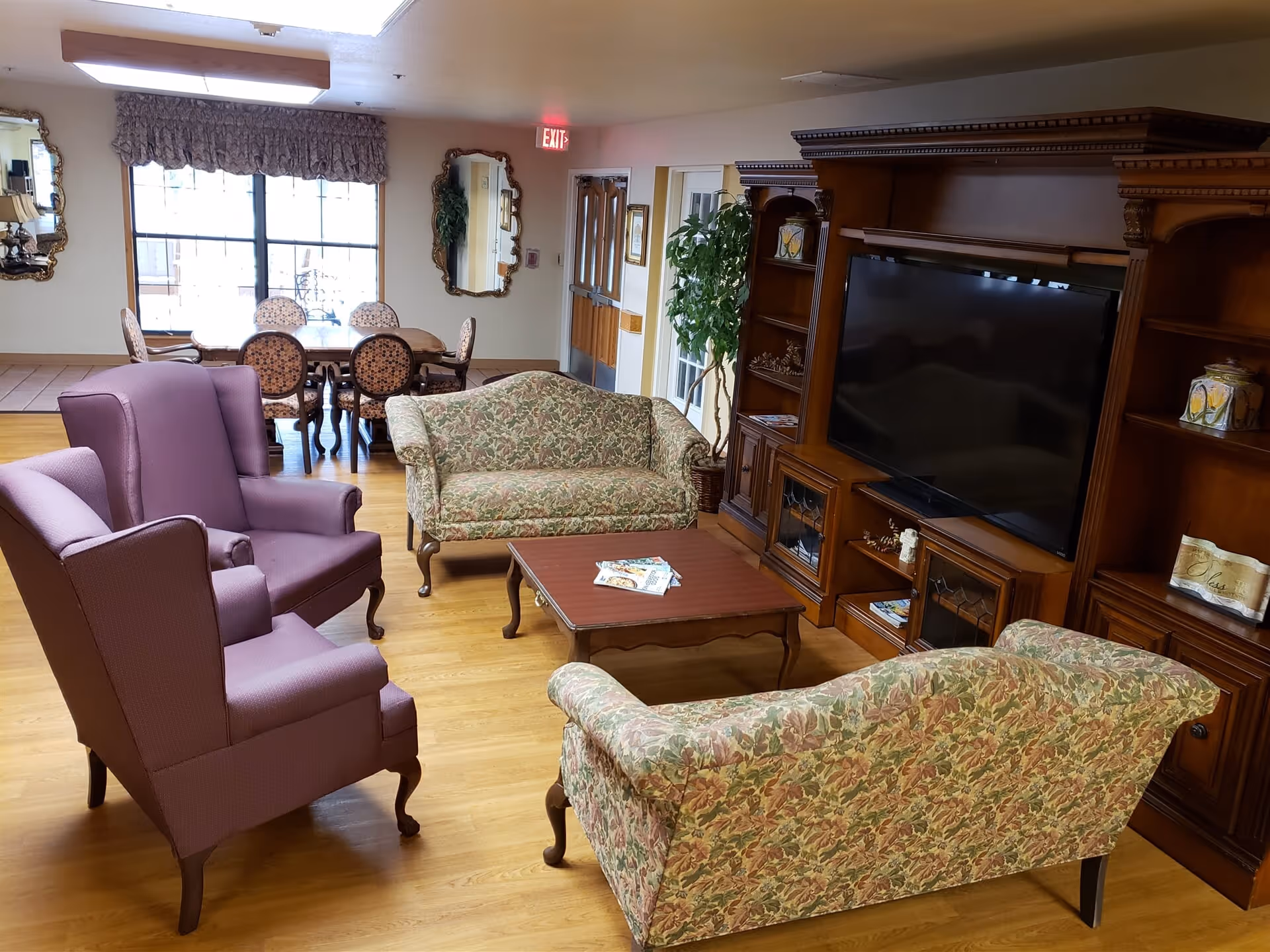 A cozy living room area in a senior living facility with two floral-patterned sofas, two purple armchairs, a wooden coffee table with magazines, and a large wooden entertainment center with a flat-screen TV. In the background, there is a dining area with a table and chairs, two decorative mirrors on the wall, a large window with a valance, and an exit door.