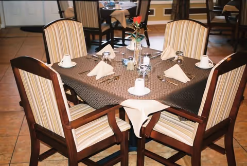 A dining table set for four with striped cushioned wooden chairs around it. The table is covered with a dark brown tablecloth and a white cloth underneath, with folded napkins, cups, silverware, salt and pepper shakers, and a small vase with a single red rose in the center. The setting is indoors with tiled flooring and other tables and chairs visible in the background.